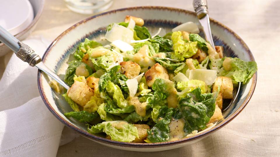 Schüssel Ceasar Salat mit Harissa Dressing​ und zwei Gabeln auf hellem Tisch mit Glasflaschen und Zitronen im Hintergrund.