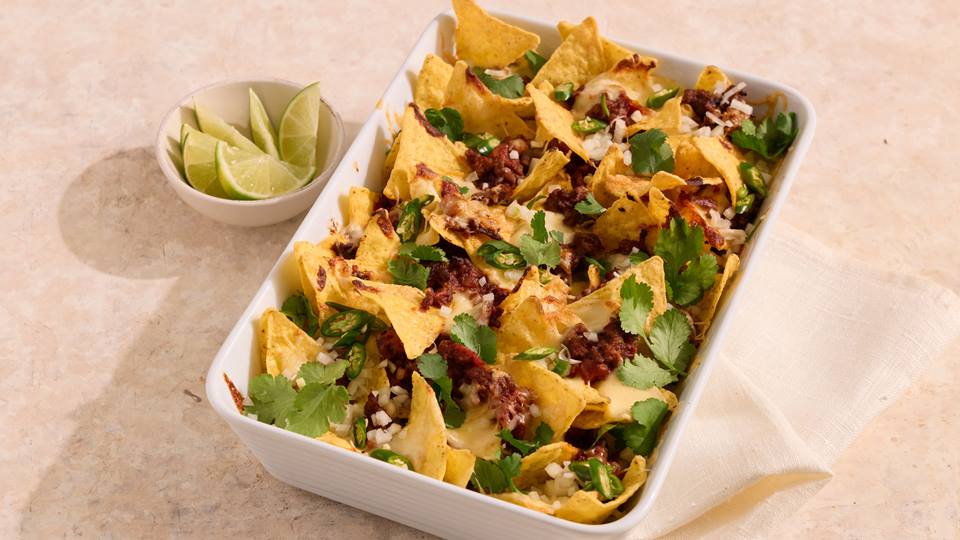 Auflaufform mit Chili-Cheese-Nachos, garniert mit Koriander und Limettenspalten daneben.