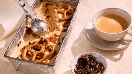 Eiscreme mit Brezeln und Karamell in einer rechteckigen Metallform neben einer Tasse Kaffee und einer Schale mit Kakaonibs