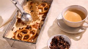 Eiscreme mit Brezeln und Karamell in einer rechteckigen Metallform neben einer Tasse Kaffee und einer Schale mit Kakaonibs