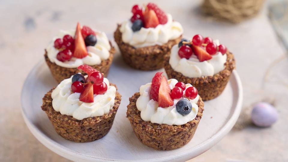 Fünf Törtchen aus Müsli mit einem Topping aus Sahne, garniert mit Heidelbeere, Erdbeere und Johannisbeere stehen auf einem Porzellanteller