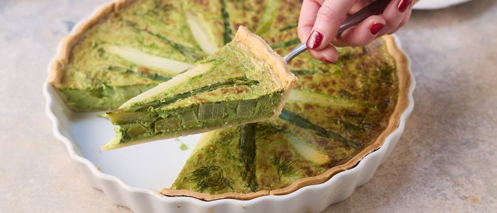 Auf einem Tortenheber wird ein Stück Spargel-Quiche gehalten, dahinter ist eine Quicheform mit der restlichen Quiche zu sehen.