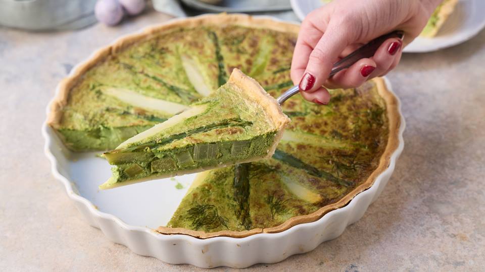 Auf einem Tortenheber wird ein Stück Spargel-Quiche gehalten, dahinter ist eine Quicheform mit der restlichen Quiche zu sehen.