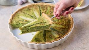 Auf einem Tortenheber wird ein Stück Spargel-Quiche gehalten, dahinter ist eine Quicheform mit der restlichen Quiche zu sehen.