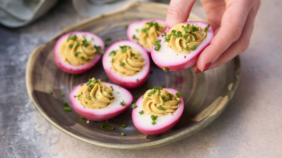 Ein halbes Ei, welches äußerlich pink gefärbt ist wird in die Kamera gehalten, dahinter sind auf einem braunen Teller weitere halbe pinke Eier zu sehen. Die Eier sind mit einer Creme und Petersilie garniert.
