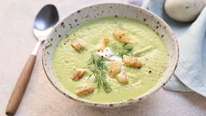 Eine Schüssel mit grüner Frühlingssuppe steht zwischen einem Esslöffel mit Holzgriff einer Stoffserviette. Die Suppe ist mit Dill und weißem Spargelköpfchen garniert.