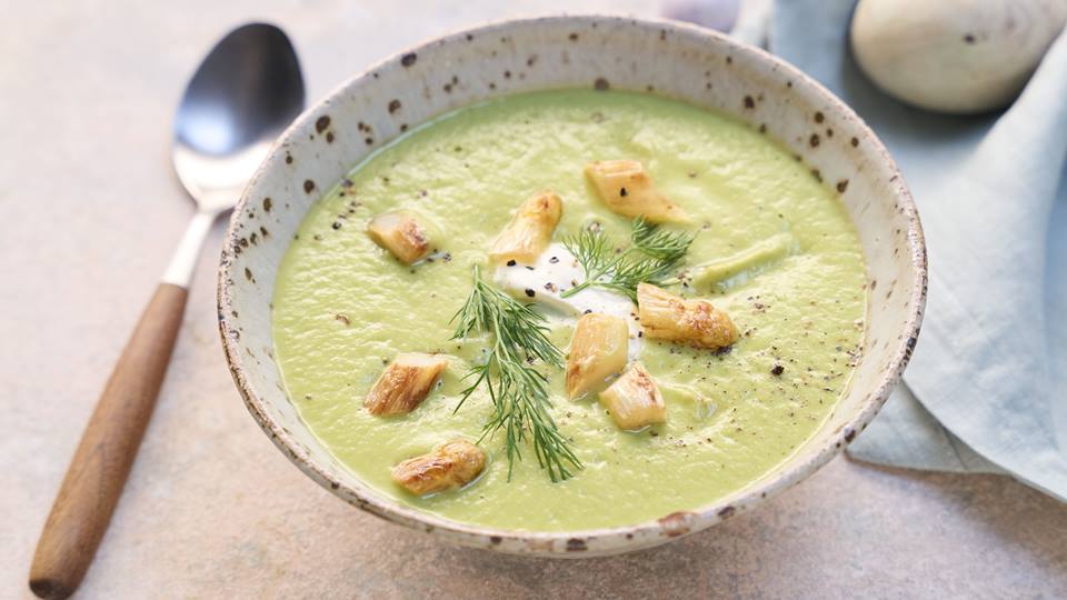 Eine Schüssel mit grüner Frühlingssuppe steht zwischen einem Esslöffel mit Holzgriff einer Stoffserviette. Die Suppe ist mit Dill und weißem Spargelköpfchen garniert.