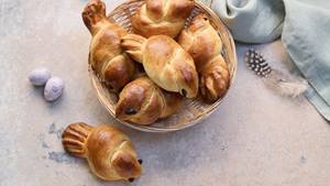 Fünf kleine Hefezöpfe, die wie Vögelchen aussehen, liegen in einem Brotkorb. Vor dem Brotkorb liegt noch ein Hefezopf-Vögelchen. Neben dem Brotkorb liegen kleine weiße Eier, eine schwarz-weiße Vogelfeder und eine hellblaue Stoffserviette.