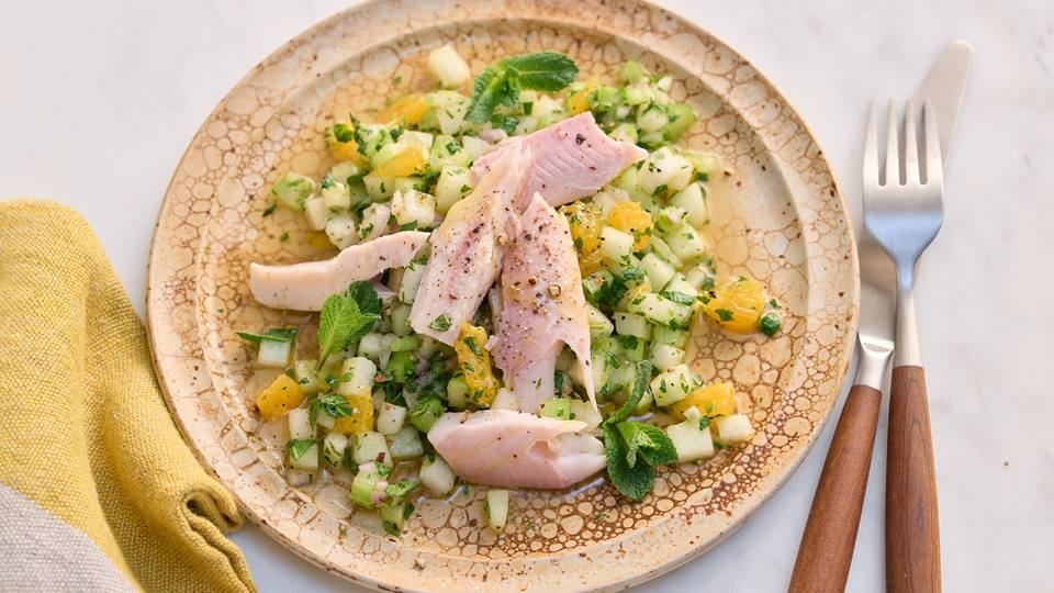 Ein Teller mit einem Gericht aus Forelle und einem fruchtigen Salat. Die Forelle liegt auf einem Salat aus Gurke, Sellerie, Orange und Minze. Der Teller ist beigefarben und hat eine leicht unebene Oberfläche. Rechts neben dem Teller liegen Gabel und Messer. Ein gelbe Stoffserviette ist links unten im Bild zu sehen. Das ganze ist auf einem hellen Hintergrund abgebildet.