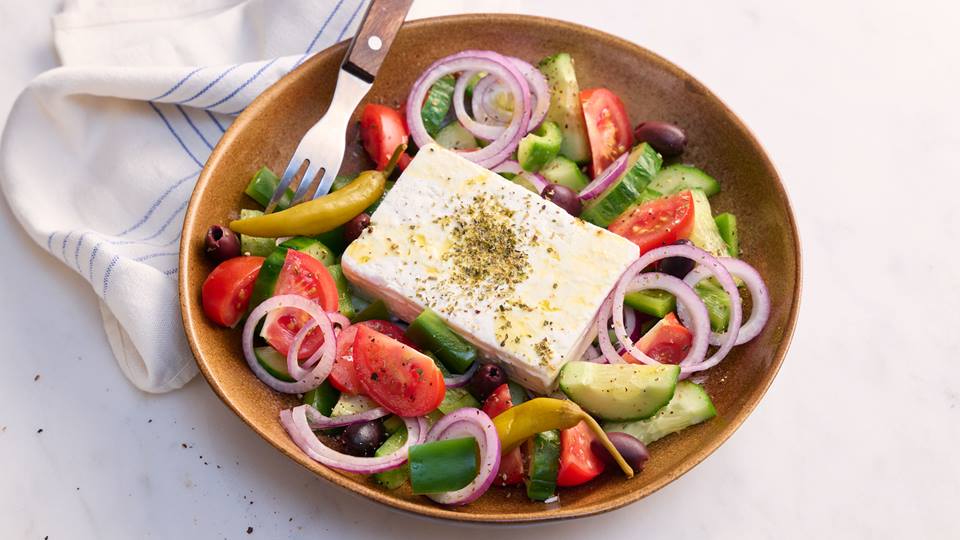 In einem braunen tiefen Teller liegt in der Mitte ein rechteckiger Schafskäse mit Gewürzen oben drauf. Im Teller neben dem Schafskäse liegen geschnittene Salatgurke, omate, Zwiebelringe, Oliven und Peperoni. Eine Gabel liegt quer im Teller und daneben eine weiße Stoffserviette mir blauen Streifen.