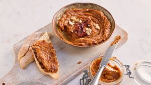 Auf einem Holzbrett steht eine kleine braune Porzellanschüssel mit Pesto rosso. Neben der Schüssel liegt ein halbes aufgeschnittenes Baguette. Auf einer Hälfte des Baguettes ist das Pesto rosso aufgestrichen. Neben dem Holzbrett steht ein Glas mit Pesto rosso auf dem ein Messer liegt.