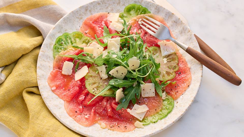 Auf einem weißen Teller liegen sehr dünn geschnittene Scheiben Wassermelone und grüne Tomate. Darüber ist Rucola und Grana Padano mit Olivenöl, Salz und Pfeffer gegeben. Rechts neben dem Teller liegt eine gelbe Stoffserviette, links auf dem Teller liegt eine Gabel mit Holzgriff und ein Messer.