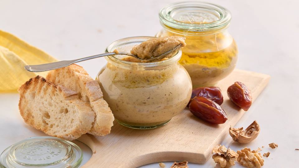 Auf einem Holzbrett stehen zwei kleine bauchige Weckgläser. Die Gläser sind mit Dattel-Walnuss-Pesto gefüllt. Ein Glas ist geöffnet und ein Teelöffel mit Walnuss-Pesto liegt darauf. Neben den Gläsern liegen Datteln, geöffnete Walnüsse und zwei Scheiben getoastete Baguettebrote. Im Hintergrund ist eine gelbe Stoffserviette zu sehen. 