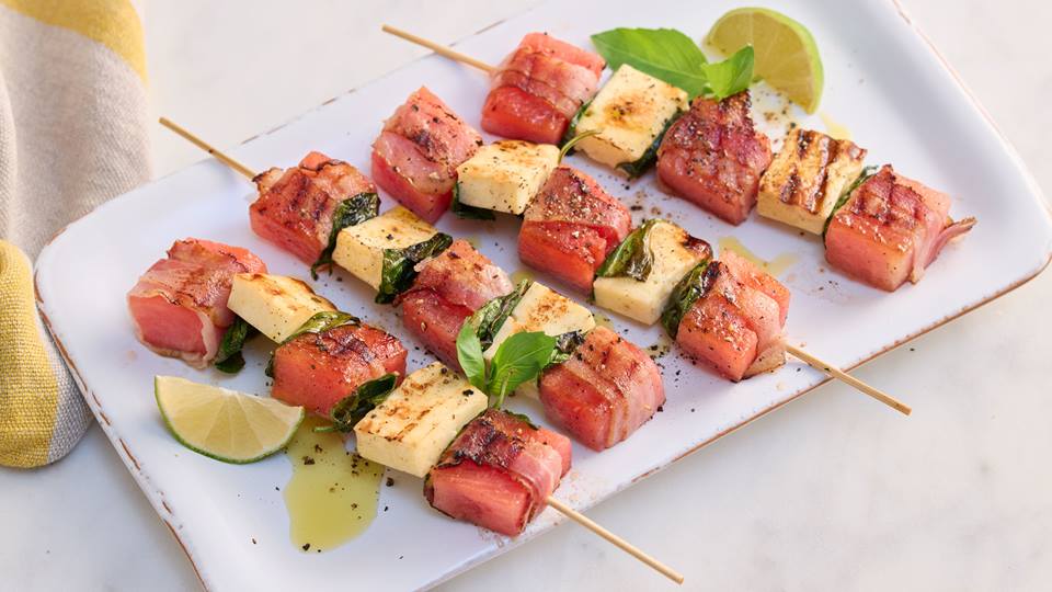 Auf einem hellen Hintergrund steht ein rechteckiges weißes Porzellantablett mit vier Wassermelonen-Bacon-Spießen und aufgeschnittenen Lemonen sowie einigen Blättern Minze.