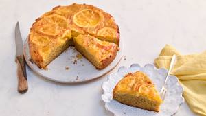 Auf einem hellen Untergrund ist ein Orangen-Kuchen auf einer weißen runden Kuchenplatte. Neben der Kuchenplatte liegt ein Messer mit Holzgriff.