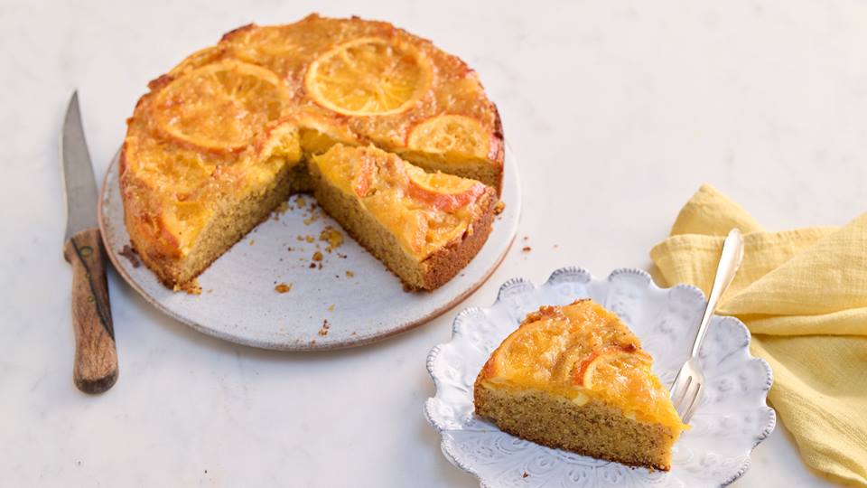Auf einem hellen Untergrund ist ein Orangen-Kuchen auf einer weißen runden Kuchenplatte. Neben der Kuchenplatte liegt ein Messer mit Holzgriff.
