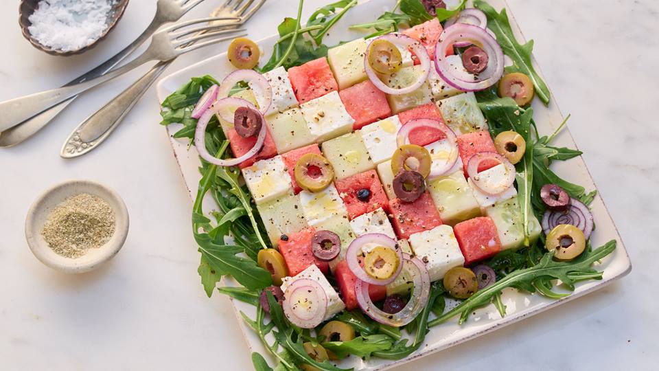 Auf hellem Untergrund ist eine weiße quadratische Porzellanplatte. Auf der Platte sind in Würfeln und als Karomuster angeordnete Stücke mit Wassermelone, Salatgurke und Feta. Der Teller ist mit Zwiebeln, Rucola, Oliven und Gewürzen garniert. Neben der Platte liegen vier Gabeln und es stehen eine Schale mit Pfeffer und eine Schale mit Salz.