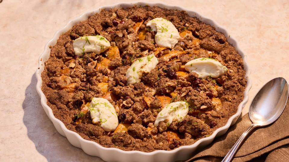 Aprikosentarte mit Streuseln und Schlagsahne-Klecksen in einer weißen, gezackten Tarteform neben einem Löffel auf einem braunen Stoff.