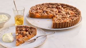 Auf hellem Untergrund steht rechts im Hintergrund auf einer weißen runden Kuchenplatte eine angeschnittene Aprikosentarte. Im Vordergrund steht ein weißer Teller mit Goldrand auf dem ein Stück Aprikosentarte, eine Kuchengabel und ein Klecks cremiger Quark ist. Im Hintergrund links steht eine kleine weiße Schüssel mit Quark und ein Glas mit einem hellen Tee, darin ein silberner verschnörkelter Teelöffel.