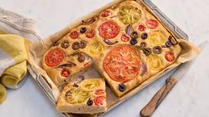 Auf einem marmorgemusterten hellen Untergrund steht eine silberne Backform mit einer Focaccia. Die Focaccia ist angeschnitten und ein Stück liegt quer in der Form. Das Focaccia ist garniert mit Tomaten, Oliven, Zwiebeln und Rosmarinzweigen. Rechts neben der Backform liegt ein Messer mit Holzgriff und links liegt ein gelb-weißes Küchenhandtuch .