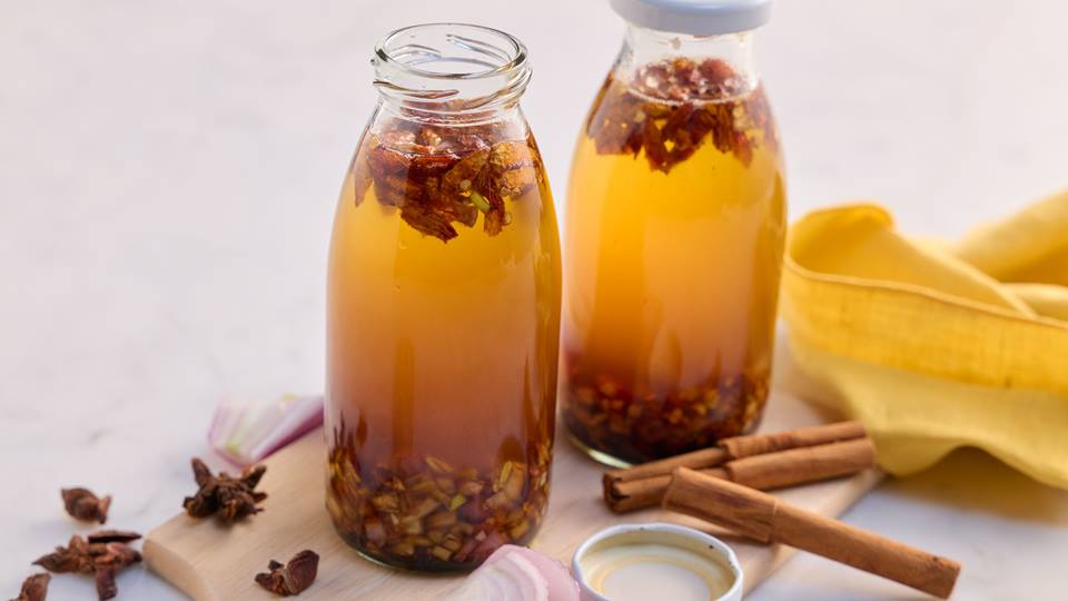 Auf einem marmorierten hellen Untergrund liegt ein Holzbrett auf dem zwei Glasflaschen mit weißem Deckel stehen. Die Flaschen sind mit Chiliöl gefüllt. In der Flasche sind kleingeschnittene rote getrocknete Peperoni zu sehen, die oben schwimmen. Unten in der Flasche sind geschnittener Knoblauch und Schalotten zu sehen. Auf dem Holzbrett neben den Flaschen liegen zwei Zimtstangen, einige Sternanis und frische Zwiebelringe. Im Hintergrund liegt eine gelbe Stoffserviette.