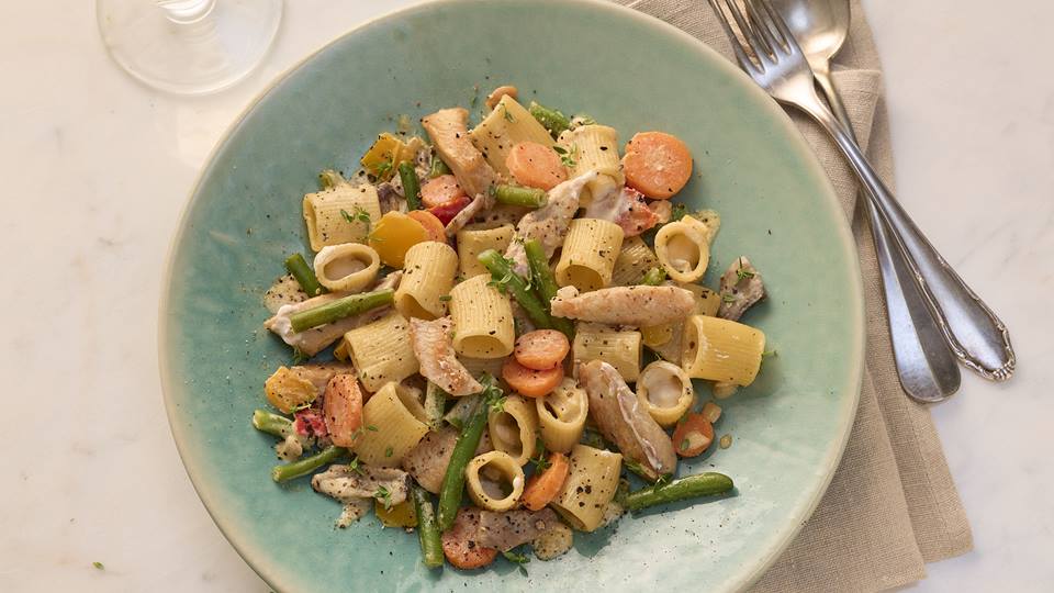 In einem tiefen türkis farbenen Teller sind Rigatoni, Bohnen, Karotten, Pilze und Putenfilet zu sehen. Neben dem Teller liegen Gabel und Esslöffel und eine braune Stoffserviette. Im Anschnitt ist ein Weinglas zu sehen.