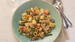 In einem tiefen türkis farbenen Teller sind Rigatoni, Bohnen, Karotten, Pilze und Putenfilet zu sehen. Neben dem Teller liegen Gabel und Esslöffel und eine braune Stoffserviette. Im Anschnitt ist ein Weinglas zu sehen.