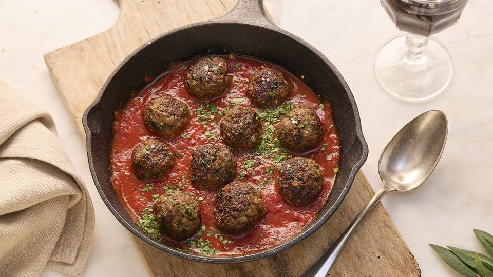 In einer gußeisernen Pfanne sind Polpette (italienische Hackfeischbällchen) in einer Tomatensauce graniert mit geschnittenen Kräutern. Die Pfanne steht auf einem Holzbret, daneben liegt ein silberner Esslöffel und eine braune Stoffserviette. Im Anschnitt ist ein Weinglas zu sehen.