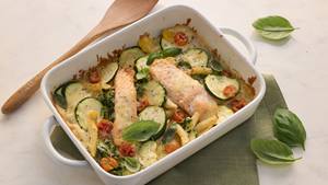 Eine weiße Auflaufform enthält mit Käse überbackenes Lachsfilet mit Zucchinischeiben, Rigatoni, Kirschtomaten und ist garniert mit Basilikum-Blättern. Die Auflaufform steht auf einer grünen Stoffserviette. Neben der Auflaufform liegt ein Holzflöffel und einige Basilikumblätter wurde verstreut.