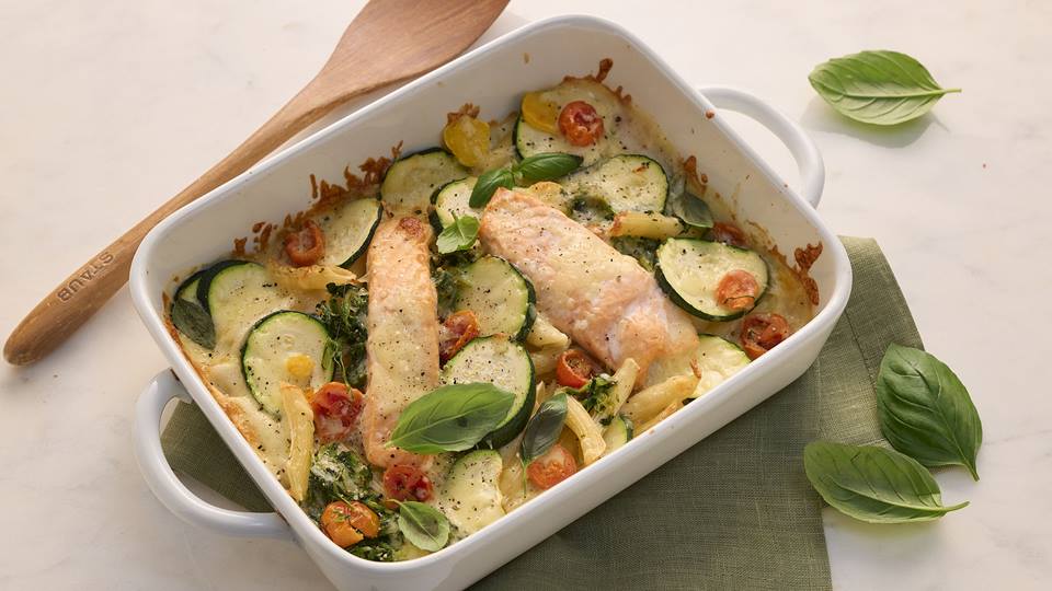 Eine weiße Auflaufform enthält mit Käse überbackenes Lachsfilet mit Zucchinischeiben, Rigatoni, Kirschtomaten und ist garniert mit Basilikum-Blättern. Die Auflaufform steht auf einer grünen Stoffserviette. Neben der Auflaufform liegt ein Holzflöffel und einige Basilikumblätter wurde verstreut.