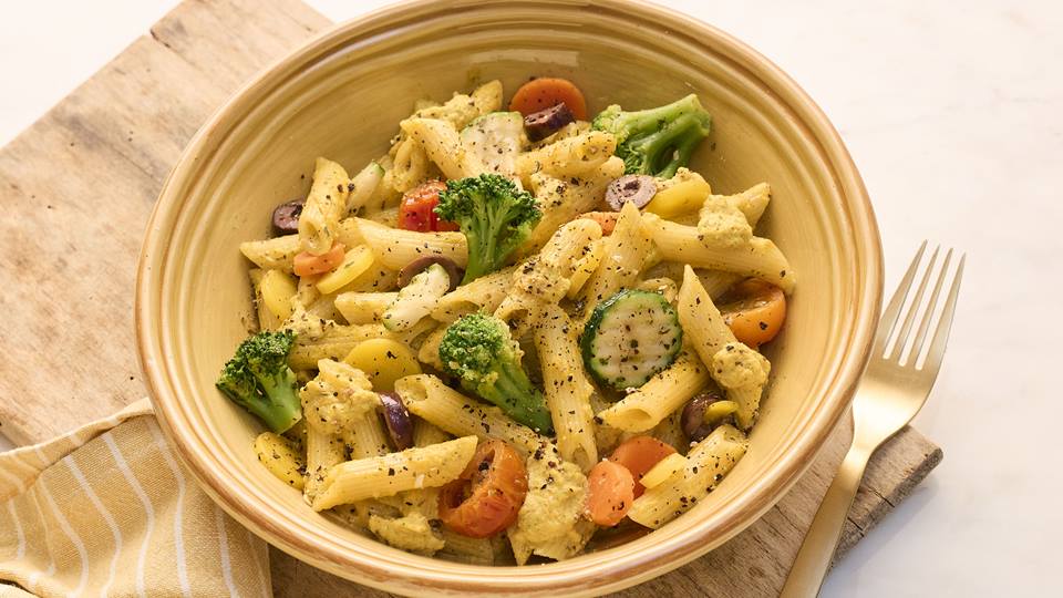 Eine Schüssel mit Penne-Pasta, gemischt mit verschiedenen buntem Gemüse wie Brokkoli, Zucchini, Karotten, Tomaten und Oliven. Die Pasta ist mit einer gelblichen Sauce überzogen und mit schwarzem Pfeffer gewürzt. Daneben liegt eine goldfarbene Gabel auf einer Serviette. Die Schüssel steht auf einem hellen Holztisch.