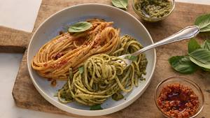 Ein flacher, weißer Teller mit zwei Portionen Spaghetti, die jeweils mit unterschiedlichen Pestos serviert werden. Links liegen die roten Spaghetti Pesto mit roten, leicht stückigen Tomaten, mit schwarzen Pfefferkörnern und Kräutern garniert. Rechts sind die grünen Spaghetti mit frischen Basilikumblättern garniert. Neben dem Teller befinden sich zwei kleine Schälchen: eines mit dem grünen Pesto und eines mit der roten Pesto. Eine silberne Gabel liegt auf dem Teller. Der Teller steht auf einem rustikalen, hellbraunen Holztisch, auf dem auch einige frische Basilikumblätter verteilt sind.