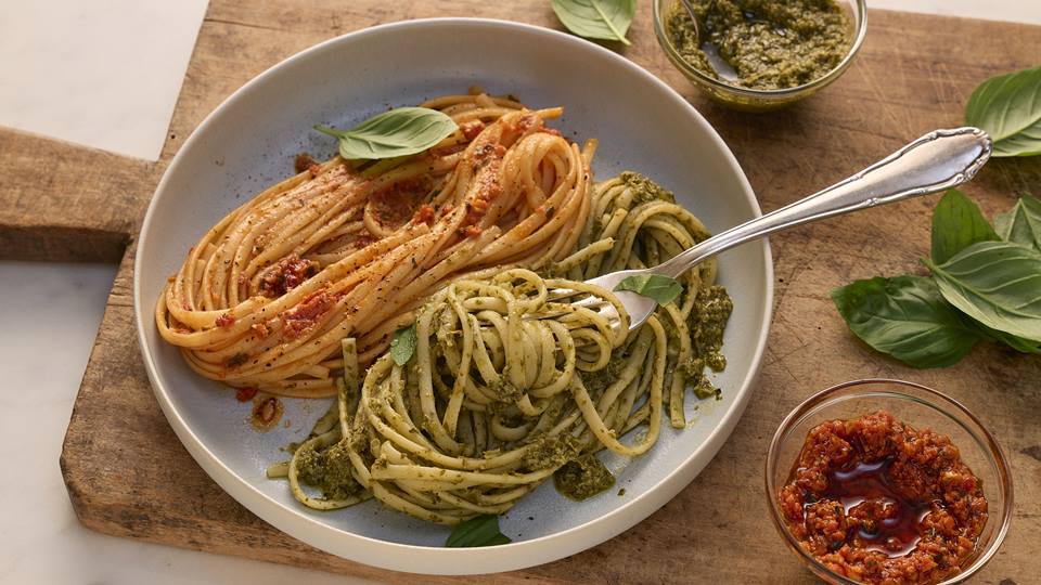 Ein flacher, weißer Teller mit zwei Portionen Spaghetti, die jeweils mit unterschiedlichen Pestos serviert werden. Links liegen die roten Spaghetti Pesto mit roten, leicht stückigen Tomaten, mit schwarzen Pfefferkörnern und Kräutern garniert. Rechts sind die grünen Spaghetti mit frischen Basilikumblättern garniert. Neben dem Teller befinden sich zwei kleine Schälchen: eines mit dem grünen Pesto und eines mit der roten Pesto. Eine silberne Gabel liegt auf dem Teller. Der Teller steht auf einem rustikalen, hellbraunen Holztisch, auf dem auch einige frische Basilikumblätter verteilt sind.