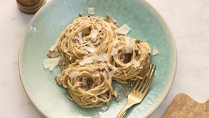 Ein Teller mit cremigen Spaghetti Cacio e pepe, garniert mit geriebenem Parmesankäse und etwas groben Pfefferkörnern. Die Spaghetti sind gleichmäßig mit der Sauce überzogen und in Nester gedreht. Der Teller ist hellblau-grün mit einem leichten Farbverlauf und steht auf einer hellen Arbeitsplatte. Daneben liegt eine goldfarbene Gabel. Im Hintergrund sind ein Stück Parmesankäse und ein Pfefferstreuer zu sehen.