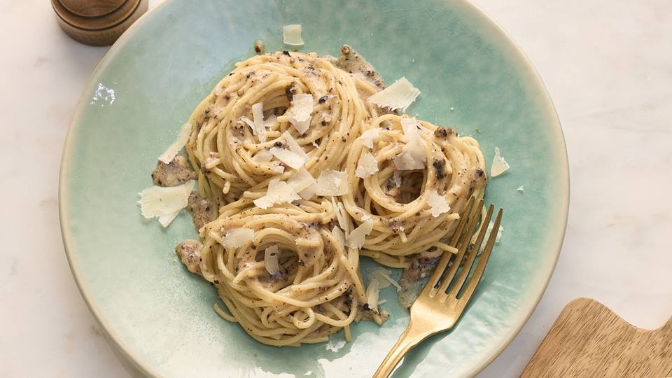 Ein Teller mit cremigen Spaghetti Cacio e pepe, garniert mit geriebenem Parmesankäse und etwas groben Pfefferkörnern. Die Spaghetti sind gleichmäßig mit der Sauce überzogen und in Nester gedreht. Der Teller ist hellblau-grün mit einem leichten Farbverlauf und steht auf einer hellen Arbeitsplatte. Daneben liegt eine goldfarbene Gabel. Im Hintergrund sind ein Stück Parmesankäse und ein Pfefferstreuer zu sehen.