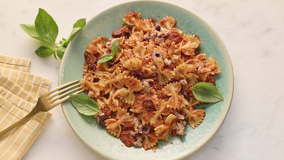 Das Bild zeigt eine Portion Farfalle-Nudeln mit Thunfisch, Oliven und einer Tomatensoße auf einem hellblauen Teller. Der Teller steht auf einer hellen Marmorfläche, und neben dem Teller liegt ein goldener Gabel auf einem gelb-weiß gestreiften Stoffuntersetzer. Ein paar Basilikumblätter sind sowohl auf dem Teller als auch daneben platziert. Das Essen sieht appetitlich und frisch aus.
