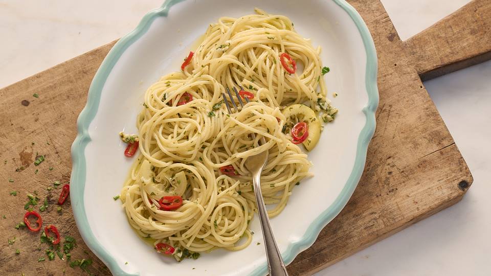 Eine Portion Spaghetti aglio e olio auf einem weißen Teller mit hellblauen Rand. Die Spaghetti sind mit Olivenöl, Knoblauch, Chili und Petersilie zubereitet und garniert. Der Teller steht auf einem alten, abgenutzten Holzbrett. Im Hintergrund ist eine helle, marmorierte Oberfläche zu sehen. Einige kleine Stücke Chili und Petersilie liegen neben dem Teller auf dem Brett. Eine Gabel steckt in den Spaghetti.Das Gesamtbild wirkt einfach, aber appetitlich und vermittelt ein Gefühl von rustikaler, mediterraner Küche.