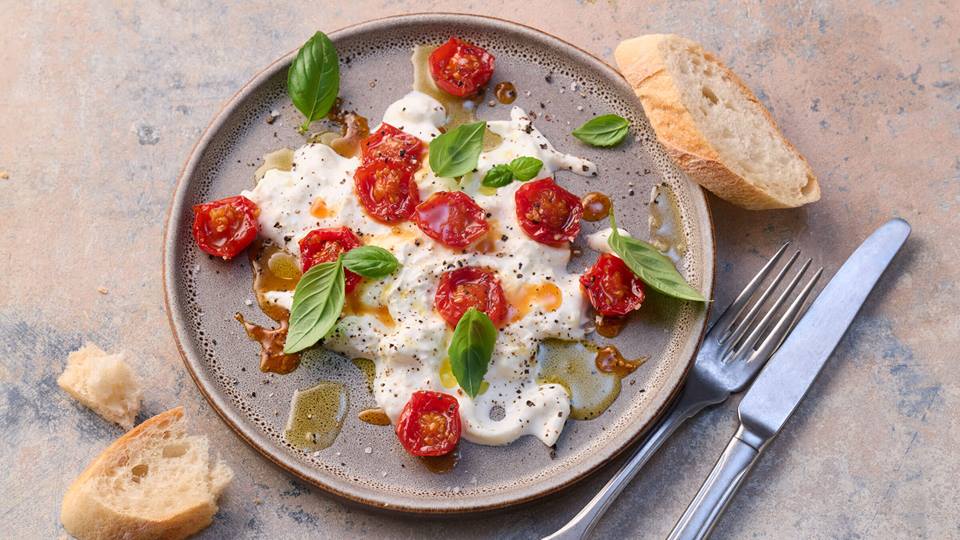 Das Bild zeigt einen Teller mit Burrata, gebratenen Kirschtomaten und Basilikum. Der Burrata ist mit Olivenöl beträufelt und mit frisch gemahlenem schwarzem Pfeffer gewürzt. Neben dem Teller liegen ein paar Brotscheiben und Besteck. Das Gericht wirkt appetitlich und einfach, aber elegant angerichtet. Der Hintergrund ist ein strukturierter, hellgrauer Untergrund.