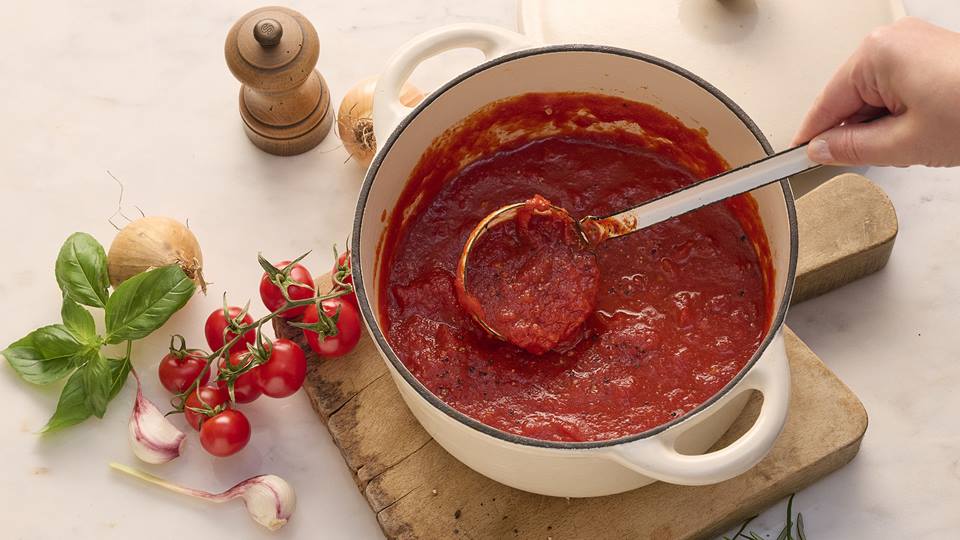 Das Bild zeigt eine weiße Schmorpfanne mit selbstgemachter Tomatensauce.  Eine Schöpfkelle mit Sauce wird herausgehoben.  Neben der Pfanne liegen auf einer Holzunterlage Cherrytomaten, Basilikum, Knoblauchzehen und eine Zwiebel. Im Hintergrund ist eine Pfeffermühle zu sehen. Das Gesamtbild wirkt appetitanregend und vermittelt ein Gefühl von hausgemachter, einfacher Küche.