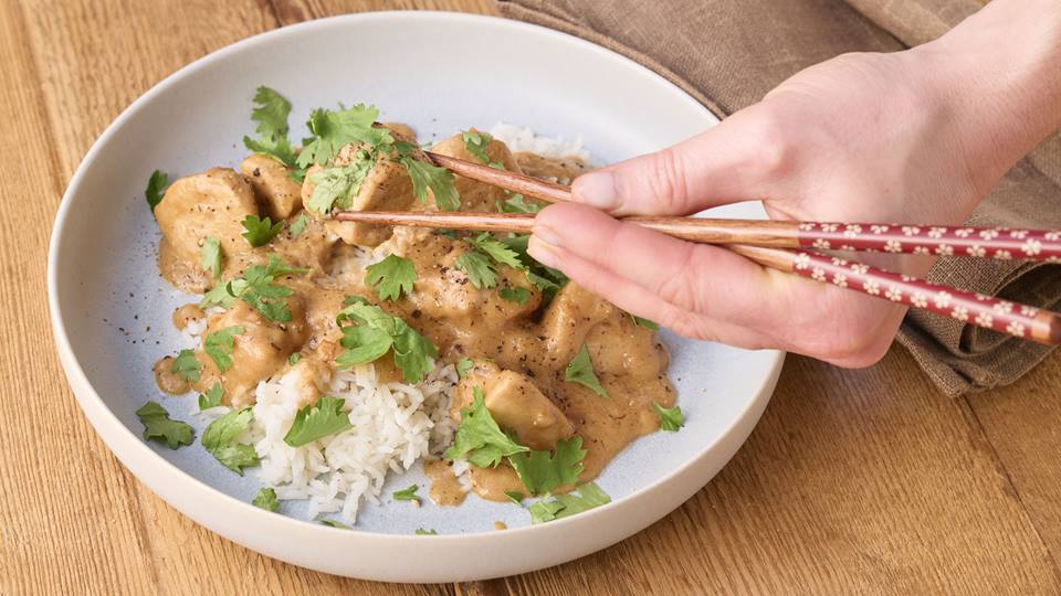 Ein weißer hoher Teller steht auf einem braunen Holzuntergrund. Im Teller sind Reis und darüber eine helle Soße mit Hähnchen-Stückchen, darüber ist glatte Petersilie gestreut. Eine Hand mit Holzstäbchen hat ein Stück Hähnchen mit Süße aufgenommen.