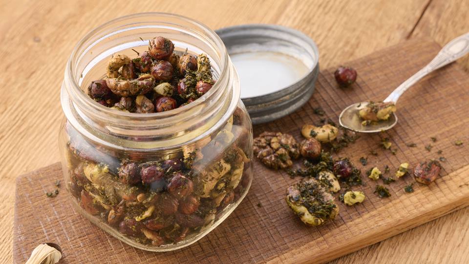 Auf einem braunen Holzbrett steht ein geöffnetes Einmachglas, der Deckel liegt daneben. In dem Glas sind unterschiedliche geröstete Nüsse, wie Walnüsse, Cashewkerne und Haselnüsse. Einige geröstete Nüsse liegen neben dem Glas auf dem Holzbrett, ebenso ein silberner Teelöffel.