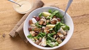 In einer weißen Porzellanschüssel ist eine Mischung aus Feldsalat, Trauben, gebratenen Champignons, Camembert-Stückchen und Weintrauben. Darüber ist eine helle Soße verteilt. In der Schüssel steckt eine Gabel. Neben der Schüssel liegt eine braune Stoffserviette und daneben steht eine kleine Glasschüssel mit der hellen Soße. In der Soße steckt ein silberner Teelöffel.