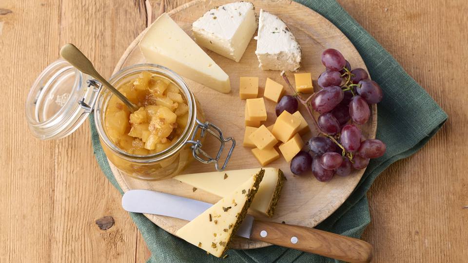Auf einem braunen Holzuntergrund steht ein Holzteller auf diesem liegen verschiedene Stücke Käse, rote Weintrauben, ein Glas mit Chutney und ein Brotmesser mit Holzgriff.  Unter dem Holzteller liegt eine grüne Stoffserviette.
