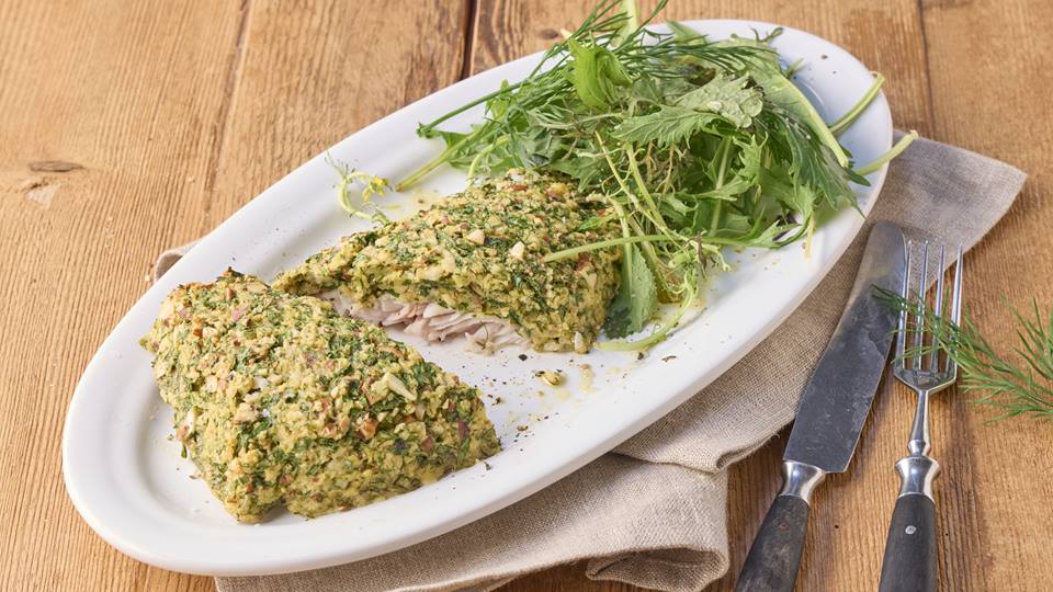 Auf einem weißen Teller ist ein Fischfilet mit Kräuterkruste und einem Salatbouquet arrangiert. Der Teller steht auf einer braunen Serviette, alles ist auf einem Holztisch. Rechts neben dem Teller liegt ein Messer und eine Gabel. ein kleiner Zweig Dill liegt auf der Gabel.