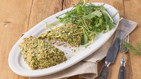 Auf einem weißen Teller ist ein Fischfilet mit Kräuterkruste und einem Salatbouquet arrangiert. Der Teller steht auf einer braunen Serviette, alles ist auf einem Holztisch. Rechts neben dem Teller liegt ein Messer und eine Gabel. ein kleiner Zweig Dill liegt auf der Gabel.