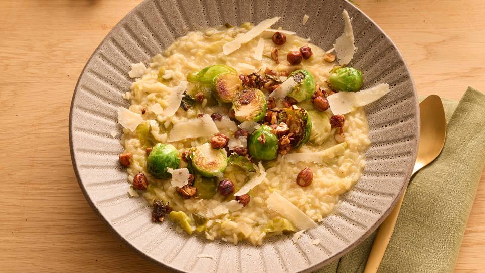 Ein Teller mit Risotto garniert mit Rosenkohl, Haselnüssen und Parmesan. Das Risotto hat eine cremige, hellgelbe Farb. Der Rosenkohl ist halbiert und auf der Außenseite leicht gebräunt. Die Haselnüsse sind ganz und geröstet, der Parmesan ist in dünnen Spänen über das Gericht gestreut. Der Teller ist aus Keramik mit einer grauen, gesprenkelten Glasur und einem gerippten Rand. Das Gericht steht auf einem Holztisch, rechts im Bild liegt eine grüne Stoffserviette.