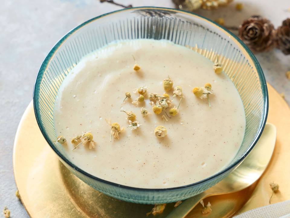 Eine Glasschale steht auf einem goldenen Teller, neben der Schale liegt ein goldener Teelöffel. In der Schale ist ein heller Vanillepudding, der mit Kamillenblüten garniert ist. Im Hintergrund sind einige Tannenzapfen zu sehen.