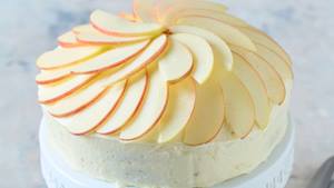 Auf einer weißen Kuchenplatte ist ein Kuchen, der mit einer hellen Creme überzogen und kunstvoll mit dünnen Apfelscheiben belegt ist. Die Apfelscheiben sind kreisförmig angeordnet und bilden eine dekorative Rosette. Der Hintergrund ist verschwommen und hell.