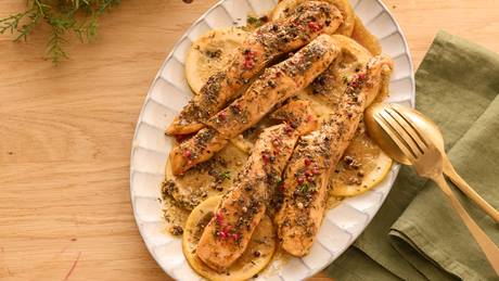 Auf einem ovalen weißen Porzellan-Teller liegen gebratene Lachsstücke, angerichtet mit Zitronenscheiben, Kräutern und Pfeffer. Neben dem Teller liegt eine grüne Stoffserviette sowie ein goldener Löffel und eine goldene Gabel.