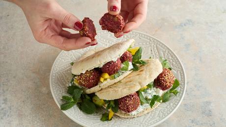 Das Bild zeigt zwei Pita-Brote, die auf einem weißen Teller liegen. Sie sind mit Falafel, Salat, Soße und frischen Kräutern gefüllt. Eine Person hält zwei weitere Falafel-Stücke über dem Teller.